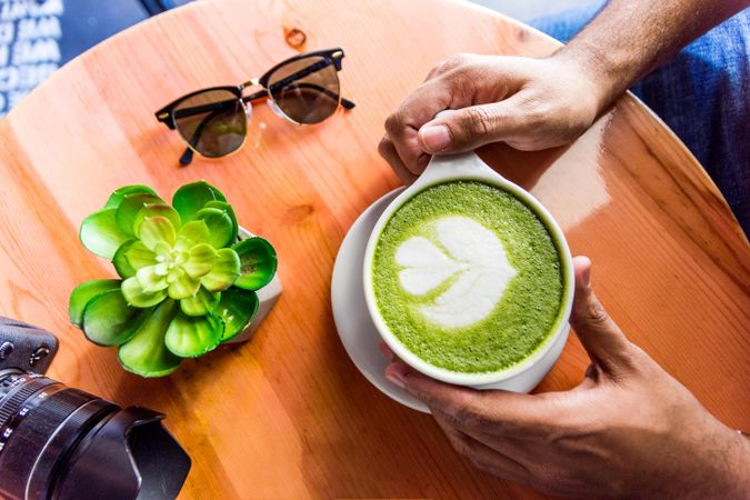 Person holding a matcha with flower shape foam beside a cactus plant and a camera and sunglasses on wooden table