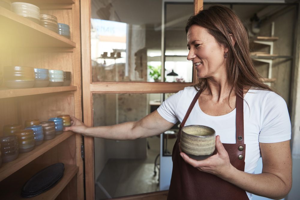 Woman in leather apron smiling while arranging handmade cups in her shop