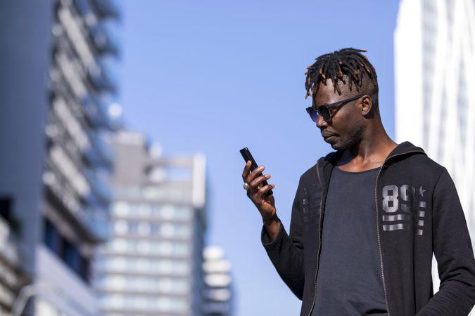 Black male with sunglasses standing against cityscape on the street while using a mobile phone in sunny day