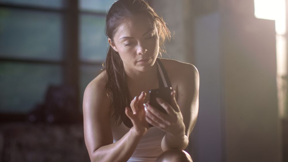 Athletic Brunette Uses Smartphone while Resting on the Bench after Her Intensive Cross Fitness Bodybuilding Gym Training.