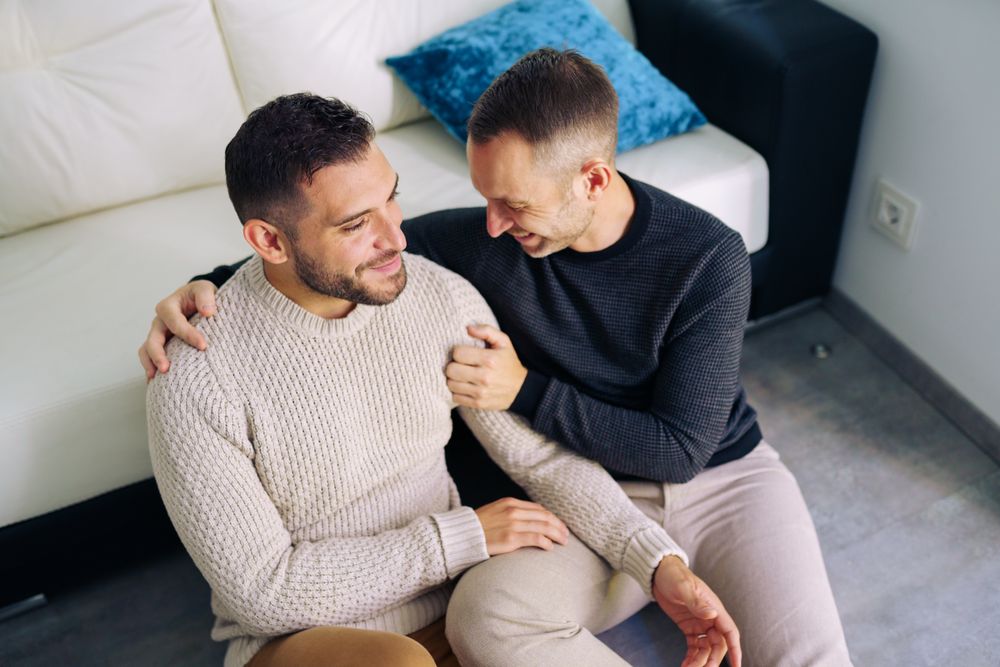 Cute male couple sitting on floor and smiling together at home