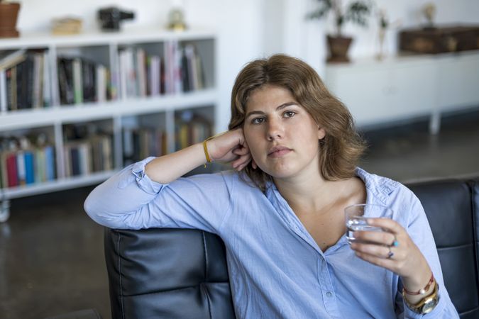 Serious young woman sitting on couch and holding glass of water