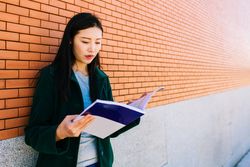 Asian student reading textbook near brick wall