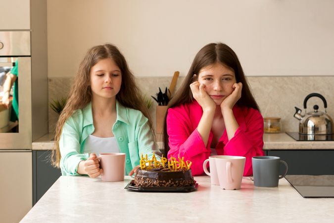 Two girls sitting with birthday cake and mugs at kitchen.