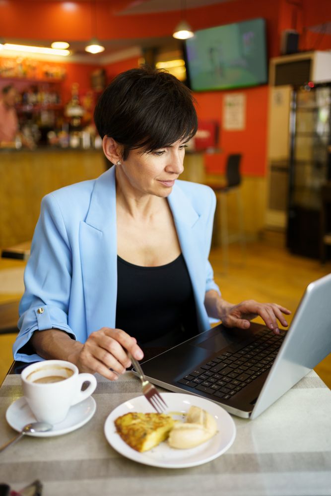 Female in trendy blue jacket sitting at table using laptop with slice of cake in cafe