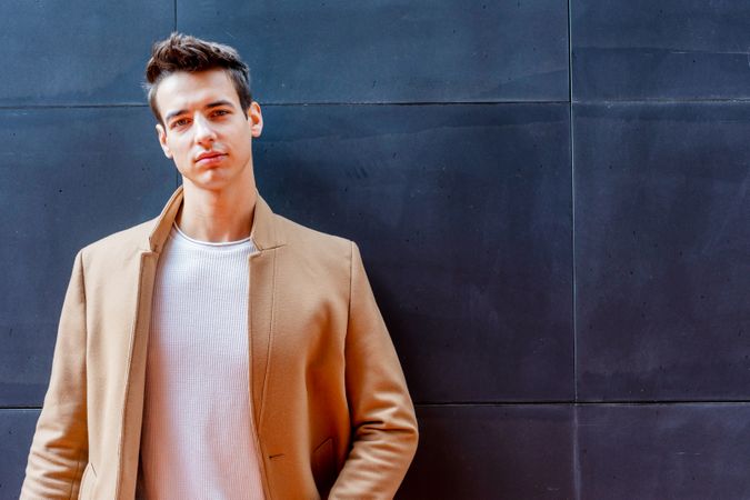 Cool male in camel coat looking up leaning against dark wall with copy space