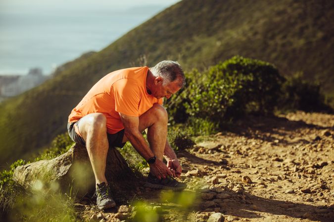 Older athletic man sitting on a rock beside a trial path and tying shoe lace