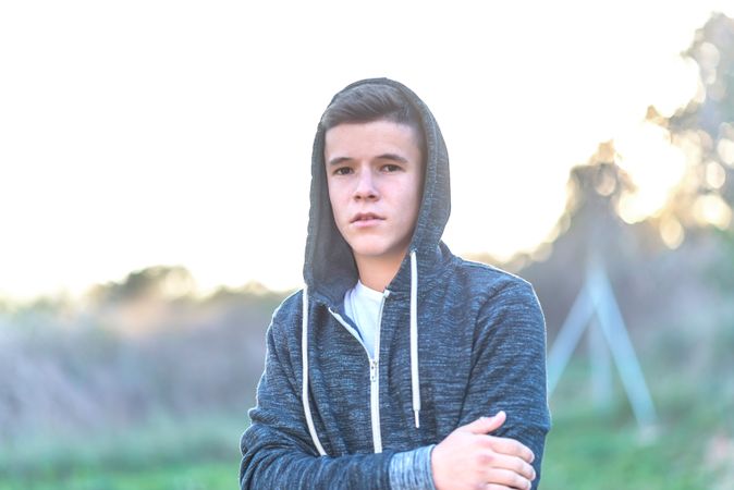 Teenage male in hoodie standing in field looking at camera with arms crossed