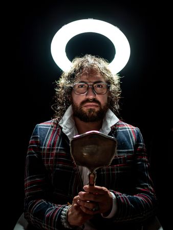Serious man with glowing fake nimbus holding mirror and looking at camera