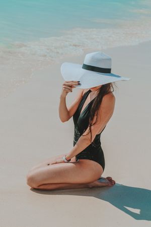 Woman in dark swimsuit wearing light sunhat hat sitting on beach