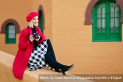 Cheerful female photographer in trendy beret and coat sitting on edge of wall with camera 41lM27
