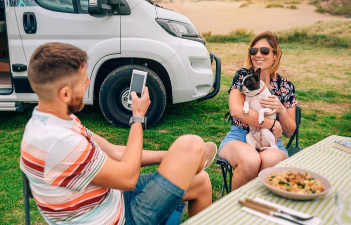 Male taking photo of smiling female sitting at table outside with cute dog