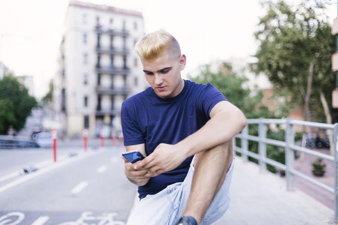 Young male with blonde hair sitting on the street while texting