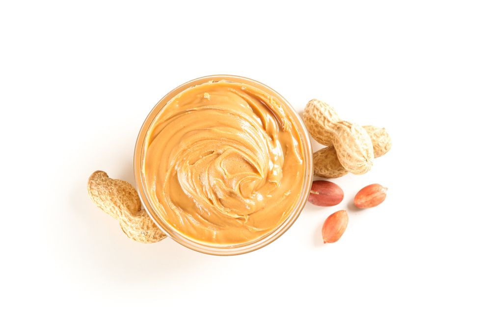 Creamy peanut butter in glass bowl and peanut isolated on light background, top view. A traditional product of American cuisine