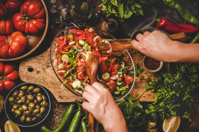 Woman tossing Mediterranean salad on wooden board surrounded with fresh vegetables and garnishes