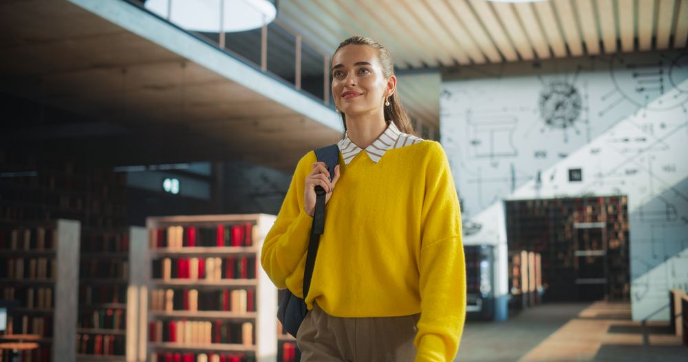 Female Student Walking in an Empty Public Library with Bookshelves. Young Smart Woman Carrying a Backpack with Textbooks to Prepare and Study for Exams. Low Angle Portrait