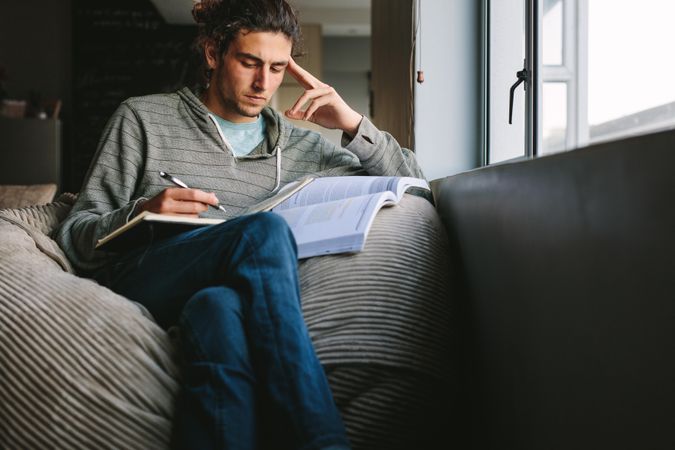Young man sitting on couch at home with books thinking deeply