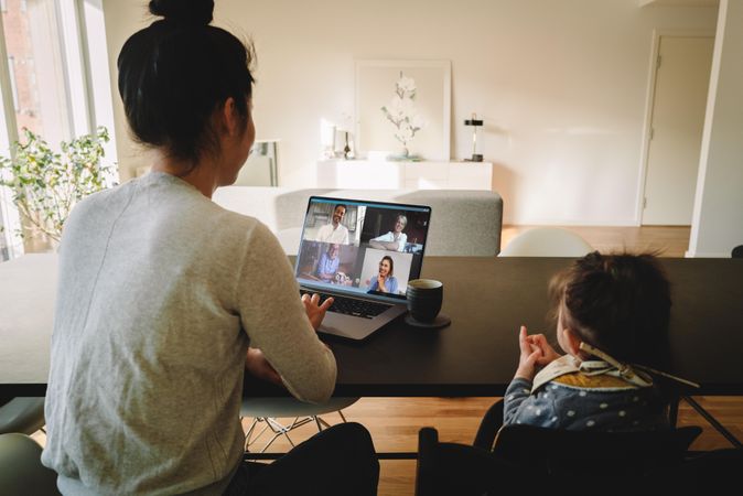 Woman working from home with her daughter sitting by at the table having a video conference call