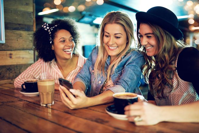Multi-ethnic group of girlfriends sharing a laugh over a cell phone at a coffee shop
