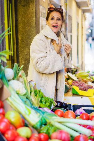 Smiling woman in warm coat standing by stall of fruits and vegetables