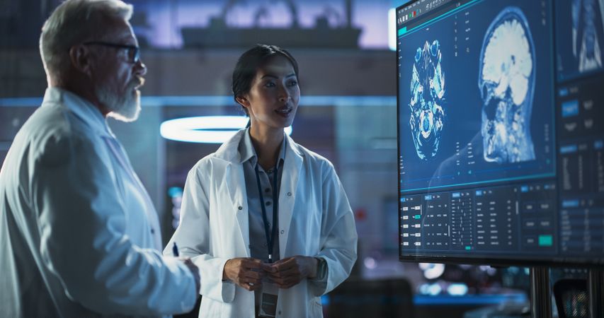 Neuroscience Laboratory Meeting: Female Asian Scientist And Male Surgeon Discussing MRI Brain Scan On TV Display.