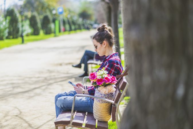 Side view of a young hipster woman sitting on a park bench relaxing in a sunny day while using a mobile phone
