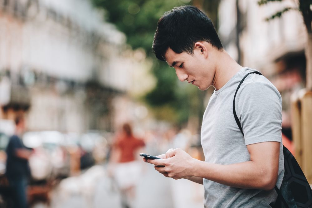 Confused Asian man using smartphone while standing on street