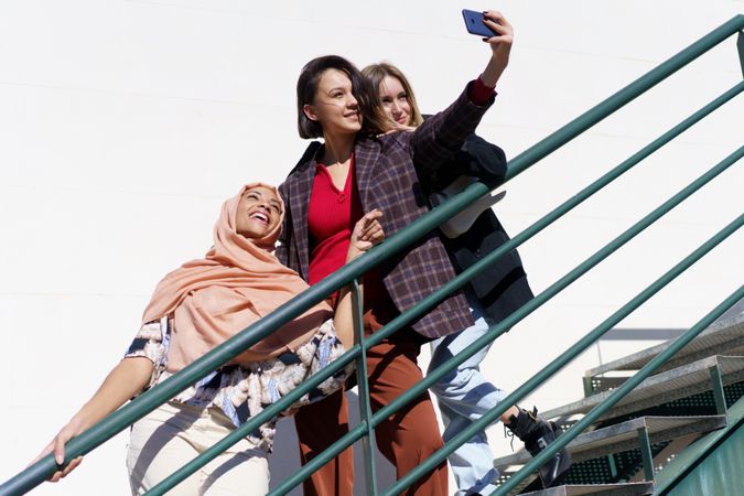 Multi-ethnic women friends, in stylish outfit and hijab taking selfie on outdoor staircase
