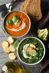 Two bowls of orange and green soup on wooden table with crackers and vegetables, vertical