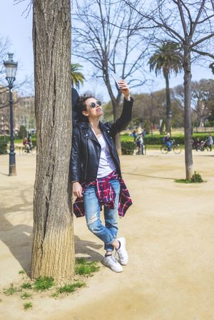Side view of a young laughing hipster woman standing and leaning on a tree at park in sunny day while using a mobile phone to take a selfie