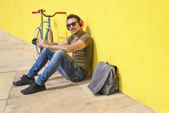 Happy male looking at camera in front of yellow wall next to bike and bag while listening to music