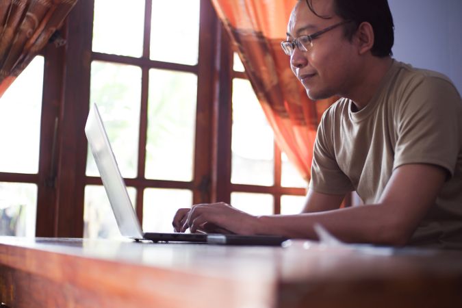 Asian Male with shirt working from home in living room with book, laptop and cup of coffee