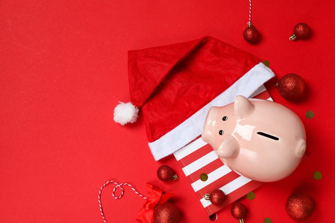 Piggy bank on gift box, balls and Santa hat on red background, space for text