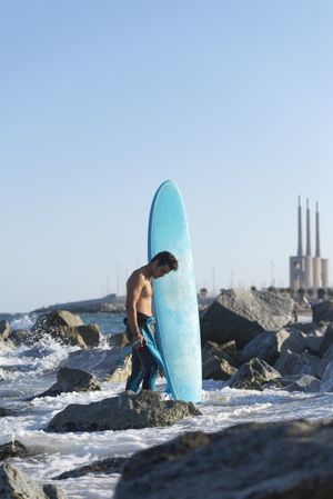 Male surfer with blue board standing in the sea around a rocky beach
