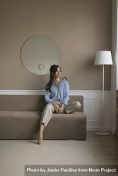Young woman sitting at the end of a brown couch in a living room while looking out the window 0v3BR5