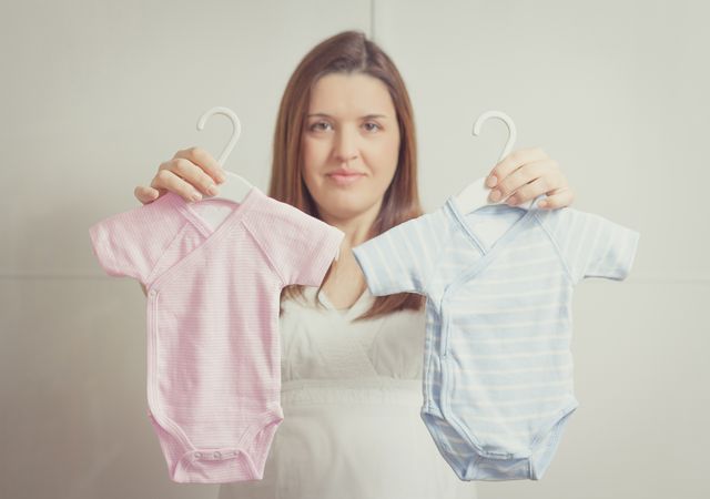 Pregnant woman holding bodysuits  for a girl or a boy in soft pastel colors
