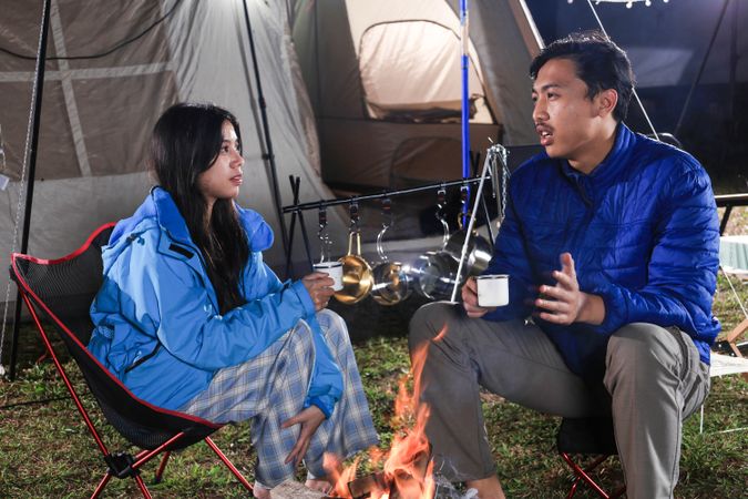 Young Asian Hetero Couple Sitting in front Tent at Campfire on Camping Site