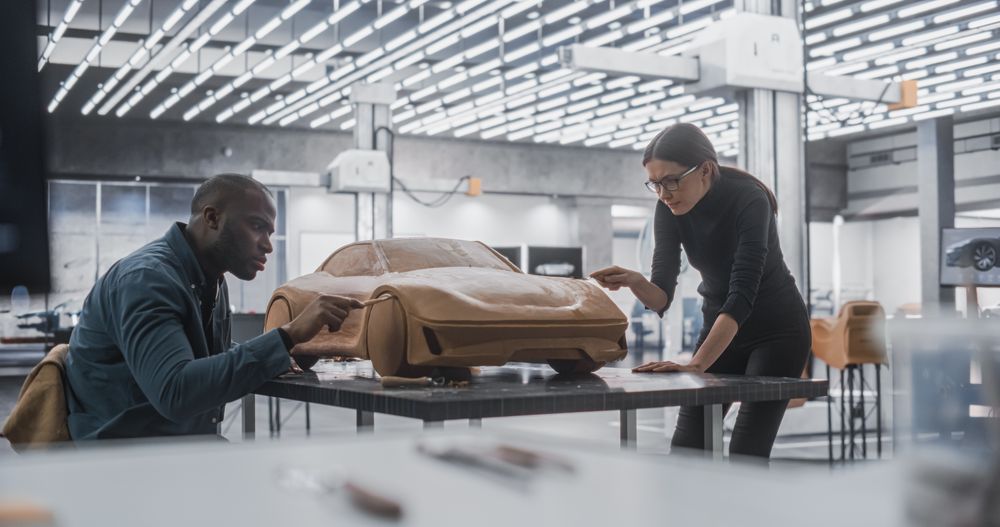 Young Female Apprentice Car Modeler Working with Black Automotive Designer on Creating a 3D Polymer Clay Model of a Modern Electric Car.