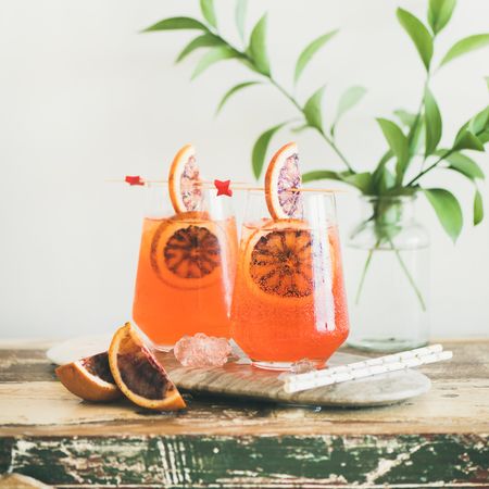 Two glasses of an orange cocktail with blood orange slices, eco straws and leaves, square crop