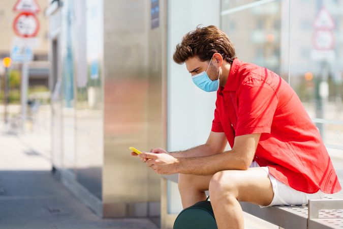 Man with facemask using his cellphone waiting for bus