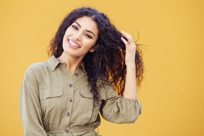 Female in army green jumpsuit smiling in front of mustard wall playing with hair