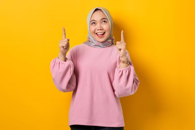 Smiling Muslim woman in scarf and pink sweater pointing up with both hands