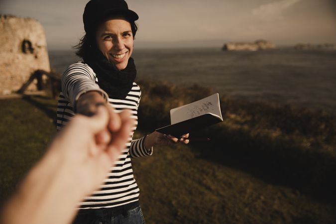 Smiling woman with notebook holding hand on shore near water