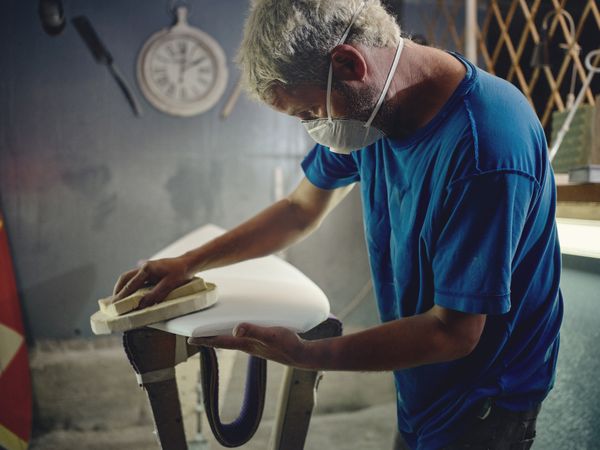 Male worker in respirator mask sanding surfboard with abrasive