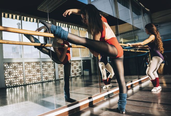 Confident young sportive women in bodysuits practicing in ballet studio stretching with barre.