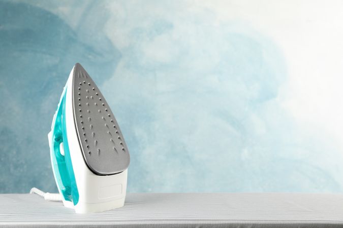 Iron on ironing board against blue background, space for text