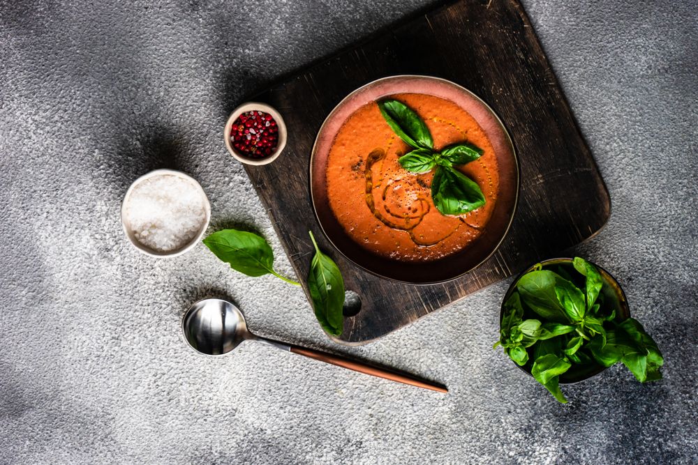 Top view of tomato soup served with oil garnish and herbs, peppers and salt