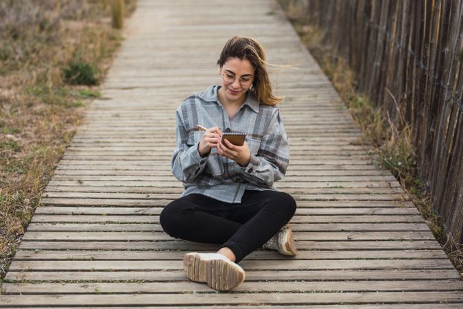 Dreamy woman sitting on boardwalk and writing in notebook in nature