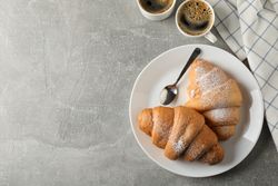 Plate with croissants, cups of coffee and towel on grey table, copy space