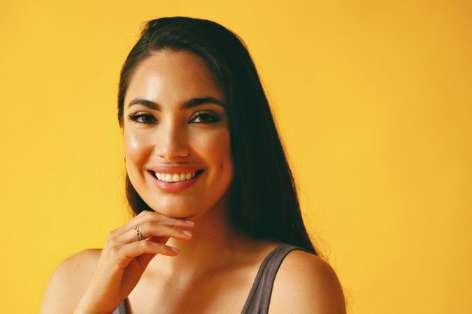 Happy Hispanic woman looking at camera in yellow room with her hand to her chin